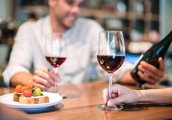 close up of a woman's hand holding a glass of red wine and a man opposite her looking at the wine bottle, smiling and holding a glass of wine too, two bruschettas on the plate