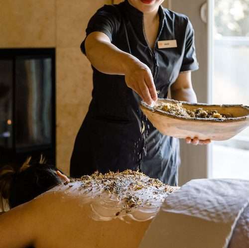 a woman receiving a body treatment at Spa Aiyana where a spa member sprinkles a spa mix onto the lotioned back