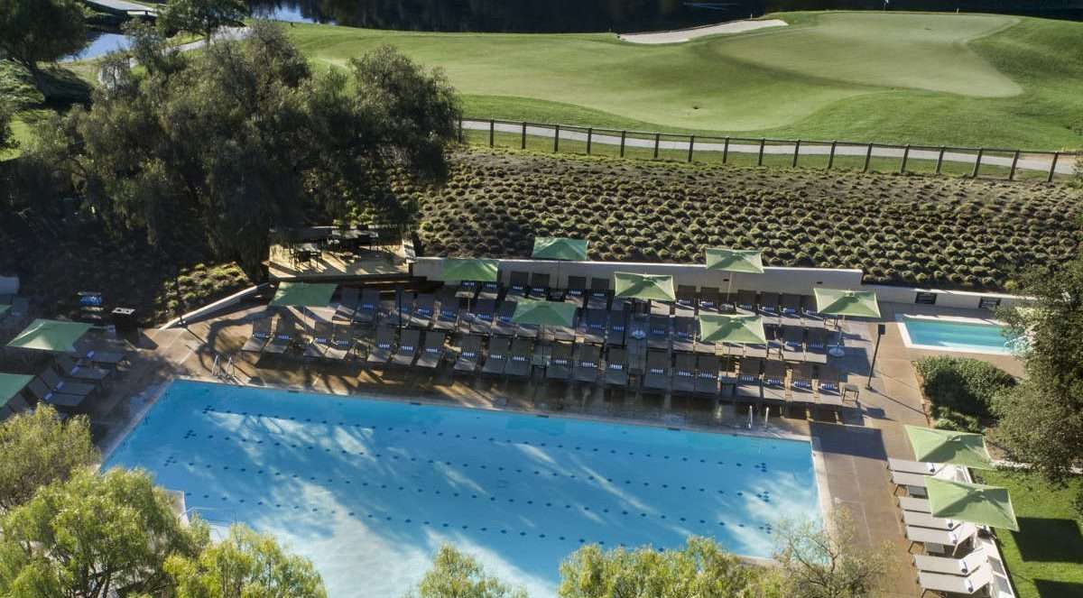 view from above of the outdoor swimming pool with mulitple grey long chairs and green sun umbrellas along the pool, view of the golf course behing the pool and green trees at Carmel Valley Ranch