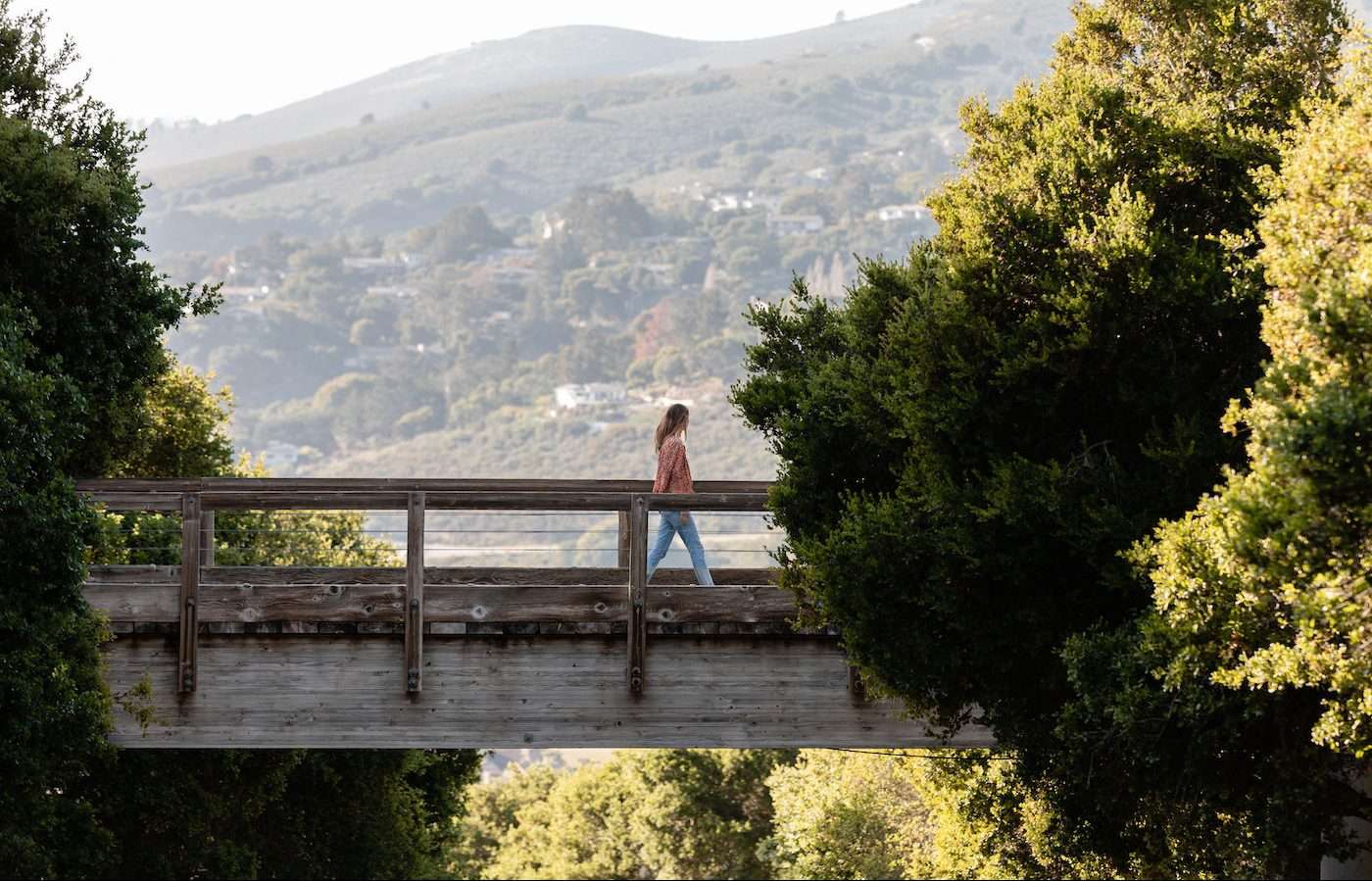 woman crossing a short high wooden bridge overlooking the valley and green trees on both side of the bridge