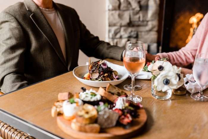Close up of a table with two dishes,a salad plate with roasted vegetables and a board with cheese and bread, a glss of rose wine and a man and a woman holding hands