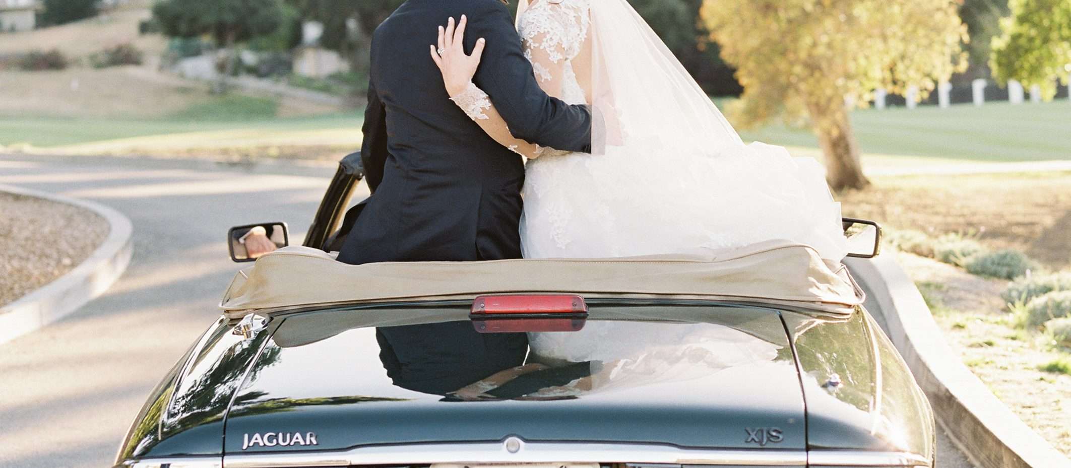Groom kissing bride's cheek as they sit on back on black car with sign that says just married